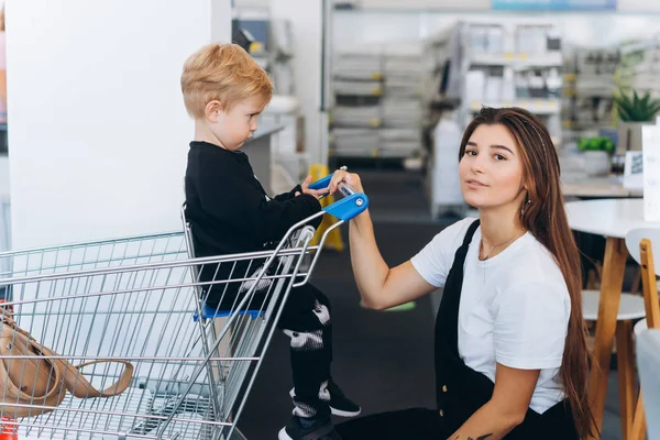 Hermosa madre lleva a su pequeño hijo en el carro del supermercado — Foto de Stock