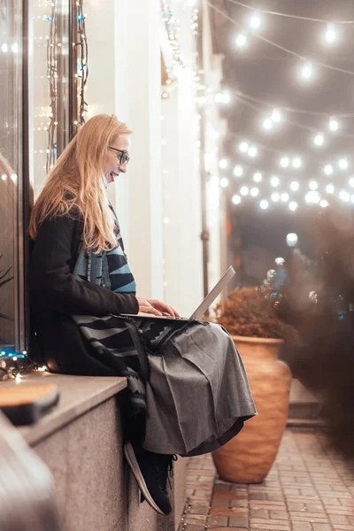 Vista lateral de la mujer joven usando el ordenador portátil en la calle con la ciudad de la noche —  Fotos de Stock