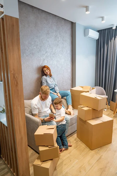 Joven familia feliz con niños desempacando cajas juntos sentados en el sofá — Foto de Stock