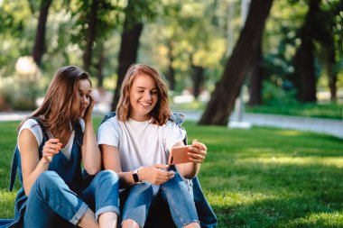 Bir çim üzerinde otururken cep telefonu tutan iki kadın arkadaş