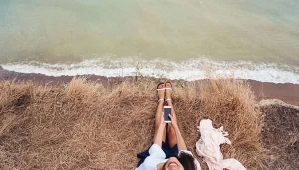 Bela jovem sentada e segurando um smartphone em suas mãos, vista superior — Fotografia de Stock