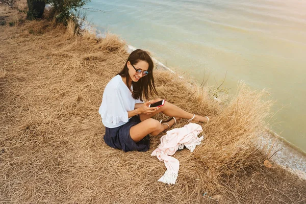 Attractive woman in summer skirt and shirt sits on the shore — Stock Photo, Image