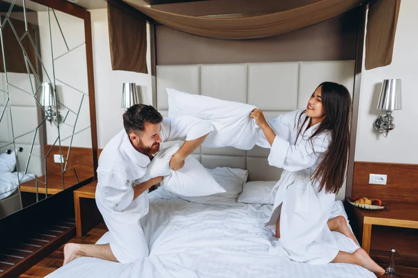 Веселая пара весело провести время в спальне борясь с большими подушками дома — стоковое фото