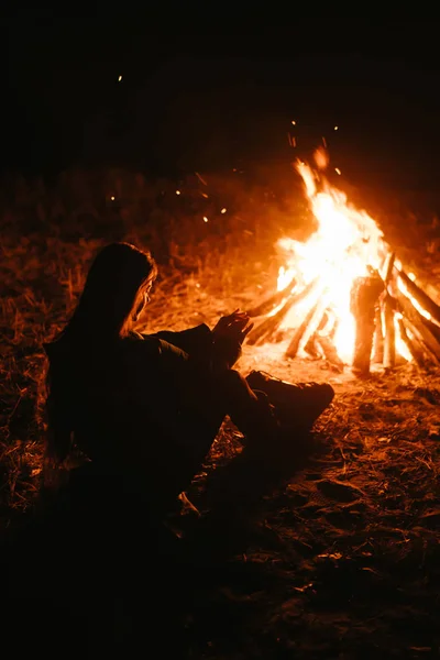 Woman sitting and getting warm near the bonfire in the night forest. Royalty Free Stock Photos