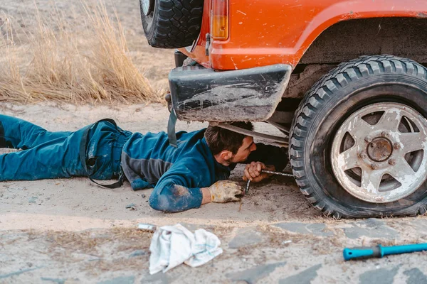 Homem encontra-se sob um carro 4x4 em uma estrada de terra — Fotografia de Stock