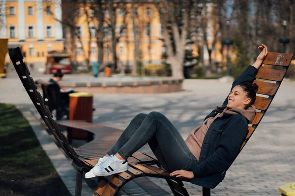 Молода жінка сидить на лавці в парку, посміхається і дивиться в камеру — стокове фото