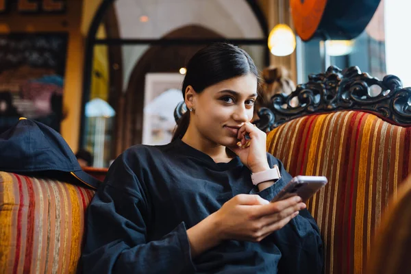 Hübsches junges Mädchen, das sich in einem Café auf einem großen weichen Stuhl ausruht und am Telefon plaudert — Stockfoto
