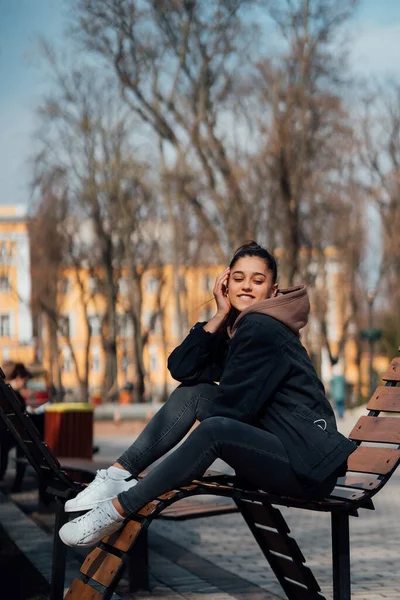 Νεαρή γυναίκα κάθεται στο παγκάκι στο πάρκο, χαμογελώντας και κοιτάζοντας στην κάμερα — Φωτογραφία Αρχείου