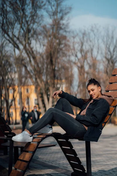 Młoda kobieta siedząca na ławce w parku, uśmiechnięta i patrząca w kamerę — Zdjęcie stockowe