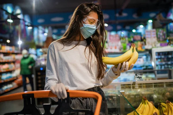 A menina com máscara cirúrgica vai comprar bananas. — Fotografia de Stock