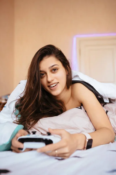 Забавная девушка лежит в постели и играет в видеоигры, держа контроллер — стоковое фото
