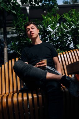 Genç adam ahşap bankta otururken elinde içecek tutuyor.