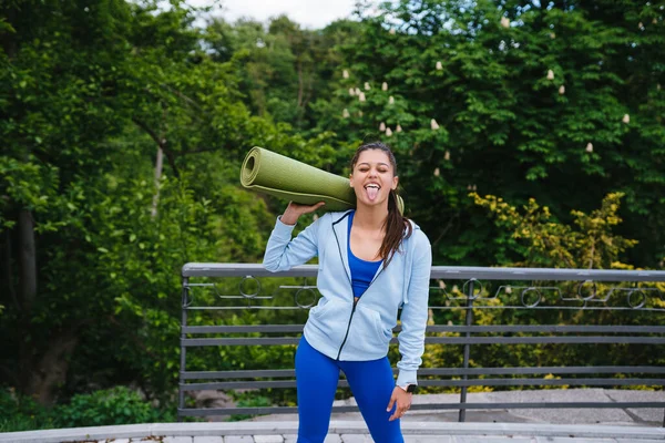 Mujer joven caminando en parque urbano sosteniendo alfombra de fitness. — Foto de Stock