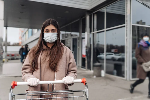 Молодая женщина в защитной маске против коронавируса 2019-nCoV толкает корзину. — стоковое фото