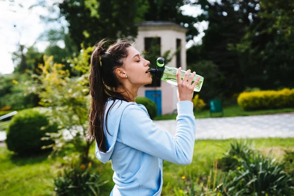 Spor takım elbiseli genç bir kadın antrenmandan sonra şişeden içiyor. — Stok fotoğraf