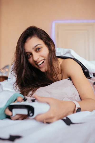 Menina engraçada deitada na cama e jogando videogame, segurando controlador — Fotografia de Stock