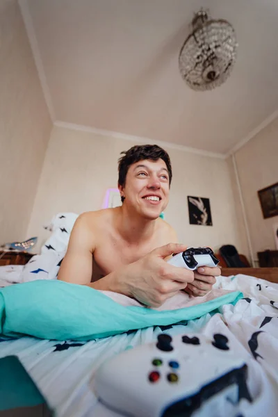 Ο τύπος ξαπλωμένος στο κρεβάτι και παίζοντας βιντεοπαιχνίδι, κρατώντας το χειριστήριο — Φωτογραφία Αρχείου