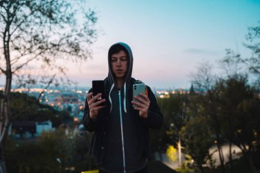Parktaki genç adam iki akıllı telefonla fotoğraf çekiyor.