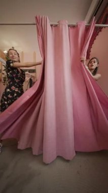 İki şık kadın parlak pembe perdelerin arkasında poz veriyor, canlı bir ortamda eğlenceli bir alışveriş deneyimi yaşıyorlar.