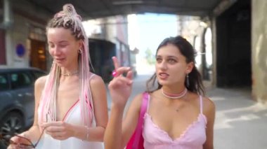 İki şık kadın hareketli bir sokakta birlikte gülüyor, moda yaz kıyafetleri ve aksesuarları sergiliyorlar.