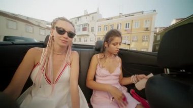 İki genç kadın neşeli bir şekilde güneşli bir günün tadını çıkarıyorlar. Üstü açık şık bir arabada, kaygısız yaz ruhlarını sergiliyorlar.