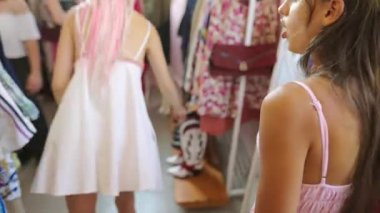 İki kız coşkuyla canlı elbiseler ve şık aksesuarlarla dolu renkli bir butiği keşfediyor.