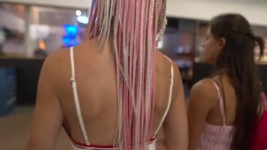 İki genç kadın, canlı ve renkli örülmüş saç şekilleriyle süslenmiş, rahat bir şekilde kapalı bir alanda geziniyorlar.