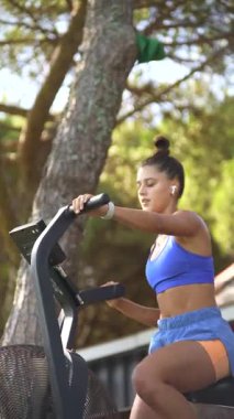 Genç bir kadın, spor yaparken eliptik bir makineye biniyor. Hem spor hem de doğa açısından güzel bir şekilde vücut buluyor.