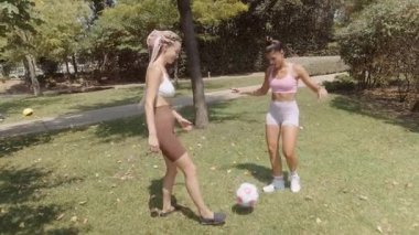 Güneşli bir park ortamında, iki enerjik kız bir futbol topunu tekmelerken keyifle eğlenirler.