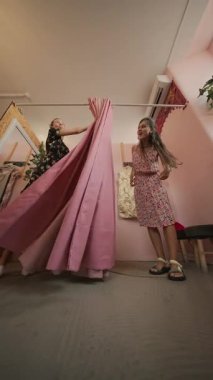 İki yakın arkadaş güzel pembe perdeleri olan şık bir odada güzel elbiseler denemekten zevk alıyorlar.