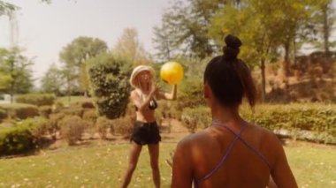 Güneşli bir günde, iki arkadaş doğayla dolu canlı bir parkta renkli bir plaj topuyla oynamaktan zevk alırlar.