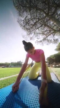 Genç bir kız hareketli bir mavi yoga minderinin üzerinde sakin bir park ortamında esneme egzersizleri yapıyor.