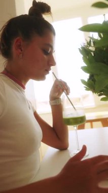 Bir kadın arkasına yaslanır ve parlak, güneşli bir odada düşüncelerine dalmış lezzetli yeşil bir smoothie 'nin tadını çıkarır.