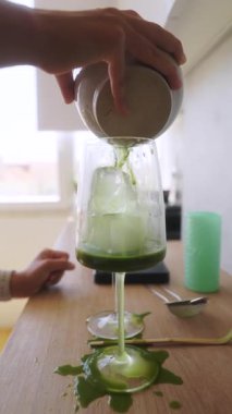Bir el, canlı yeşil bir içeceği bir bardağa ustalıkla doldurur, canlı ve davetkar bir mutfak atmosferi yaratır.