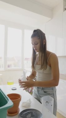 Genç bir kadın, açık ve neşeli bir mutfak ortamında ev bitkileri için saksı toprağı hazırlıyor.