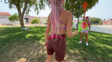 İki arkadaş heyecanlı bir şekilde dışarıda raket oynuyorlar, renkli ekipmanlar kullanıyorlar ve güneşin tadını çıkarıyorlar.