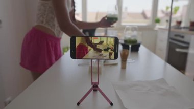 Parlak bir mutfakta sağlıklı bir içecek hazırlayan bir kadın, akıllı telefonunu birlikte yemek pişirmek için kullanır..