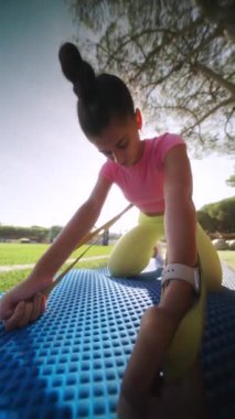 Genç bir kız açık havada esneme egzersizi yaparken canlı mavi bir paspas üzerinde direnç bandı kullanıyor.