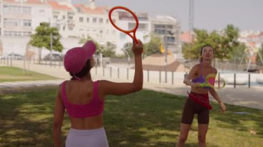 İki kadın, güneşli bir parkta raket oynayarak, dostluklarını ve spor yolculuklarını sergileyerek eğlenceli bir gün geçiriyorlar.