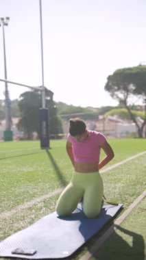 Bir kadın direniş bantlarını kullanırken açık hava ortamında spor minderinin üzerinde diz çöküyor.