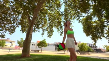 Güneşli bir parkta neşeli bir kadın renkli nesnelerle hokkabazlık yapar, çevikliğini ve dengesini göstererek eğlenir.