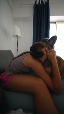 Bir kız, sevimli Fransız bulldoguyla sıcak bir kanepede otururken sevgi dolu anların tadını çıkarıyor.