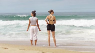 Uçsuz bucaksız okyanusun kenarında neşeyle duran iki kadın kumsalın ve dalgaların çarpıcı güzelliğini tamamen kucaklıyor.