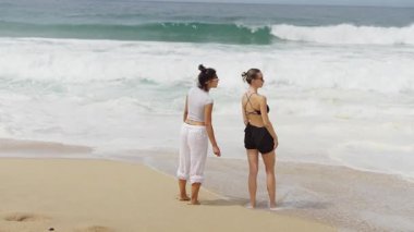 İki neşeli kadın güneşli sahil atmosferinde eğleniyor, neşeyle dalgaları kucaklıyor ve şık mayoların sıcaklığını kucaklıyor.