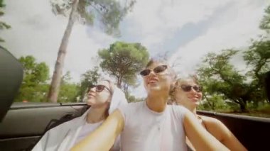 Üç yakın arkadaş neşeyle gülüyor ve açık mavi gökyüzünün altında üstü açık şık bir arabada güneşleniyorlar.