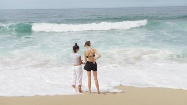 Sıcak ve güneşli bir plaj gününde, iki kadın kıyıda yan yana durur, okyanus dalgalarının tadını çıkarırlar.