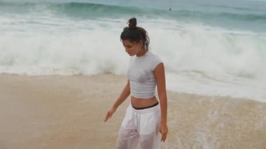 Sahilde duran genç bir kadın, dalgalar arka planda çarpıyor, sakin bir sahil havası yayıyor..