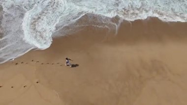 Sakin bir hava manzarası, kumlu bir plajda, yumuşak dalgalarla gezinen bir insanın rahatlatıcı bir atmosfer yarattığını gösteriyor.