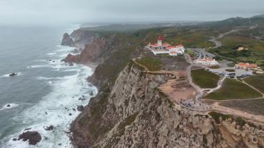 Bir hava manzarası okyanus dalgalarına karşı bir uçurum kenarı feneri sergiliyor, doğanın ham enerjisini vurguluyor.