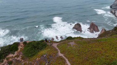 Hava manzarası dalgaların kayalara çarptığı engebeli bir kıyı şeridini ve keşfedilmesi gereken manzaralı bir patikayı gösteriyor.